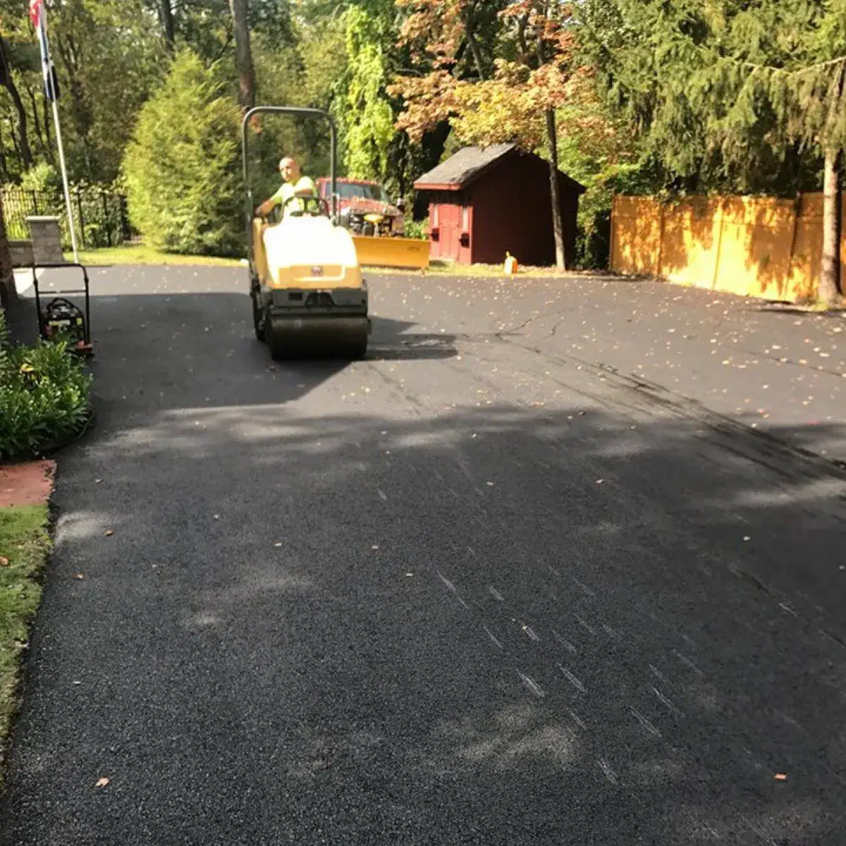 A person operates a yellow road roller to smooth freshly laid asphalt in a residential driveway surrounded by trees, a red shed, and a yellow fence.