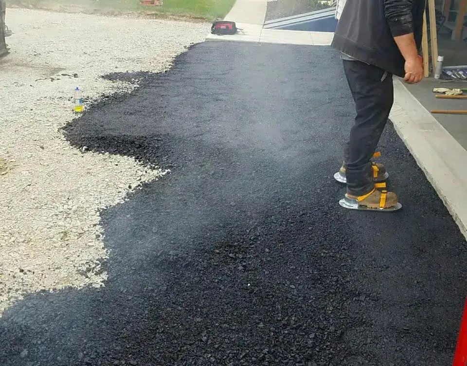 A person using metal-soled shoes stands on freshly laid, still steaming asphalt next to a gravel area, smoothing the surface near a building entrance.