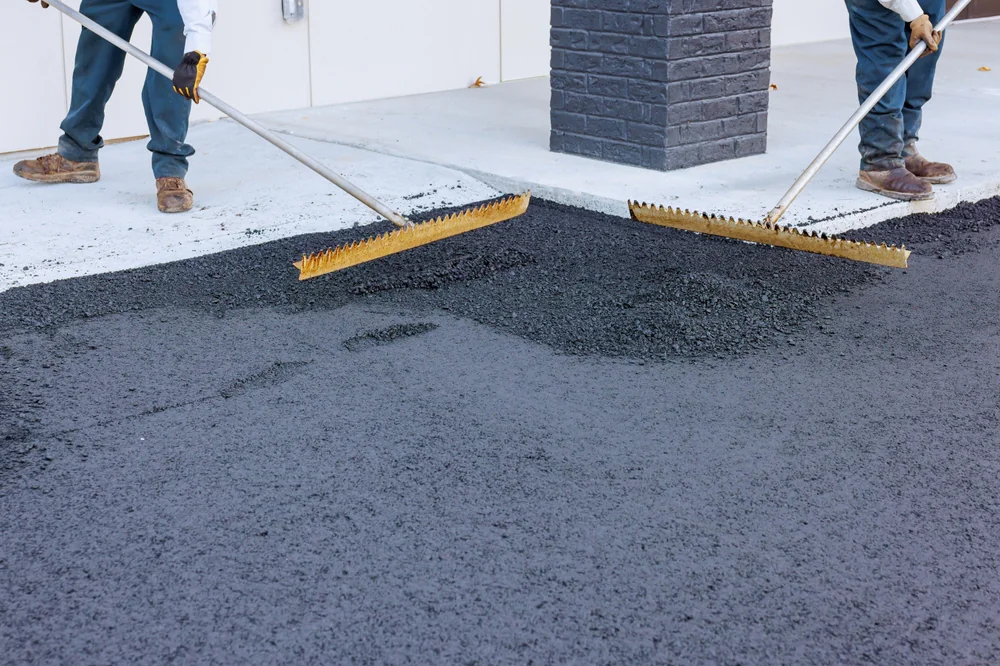 Two workers from a paving contractor Saratoga & Warren County use large rakes to spread fresh asphalt on a driveway near a building with a brick pillar in NY.