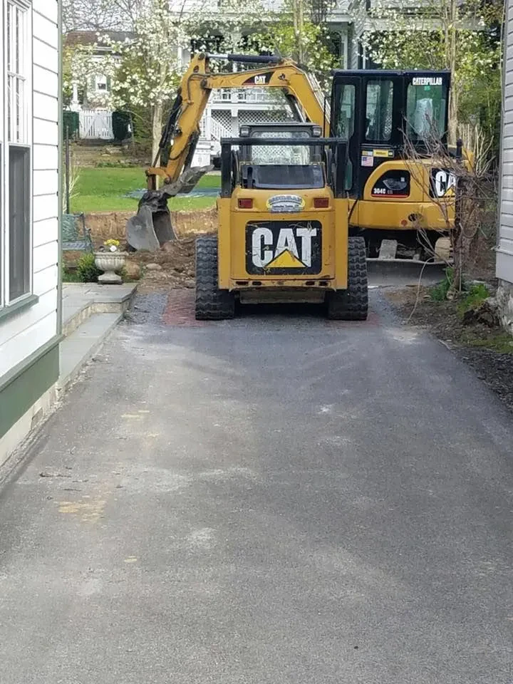 A small Caterpillar skid steer and a larger Caterpillar backhoe, used by a paving contractor Saratoga & Warren County, are parked on a driveway between two houses in NY, with blooming trees and green grass in the background.