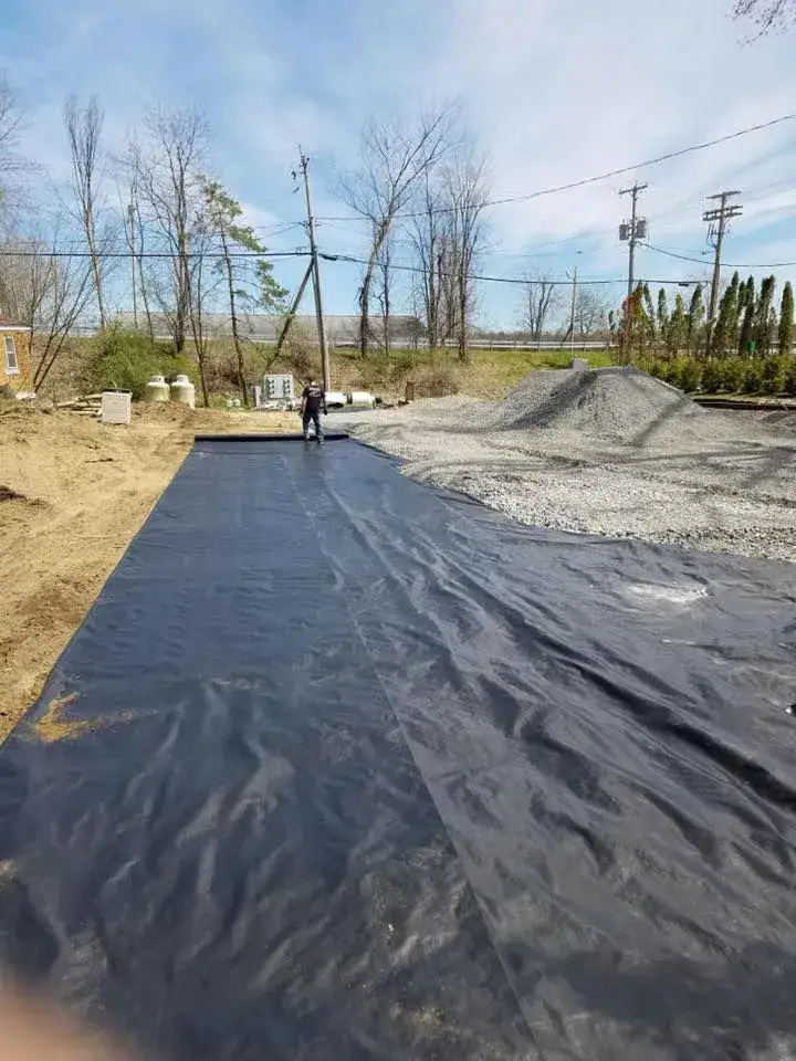 A person spreads a large black plastic tarp over the ground at a construction site, with piles of gravel and some bare trees in the background.