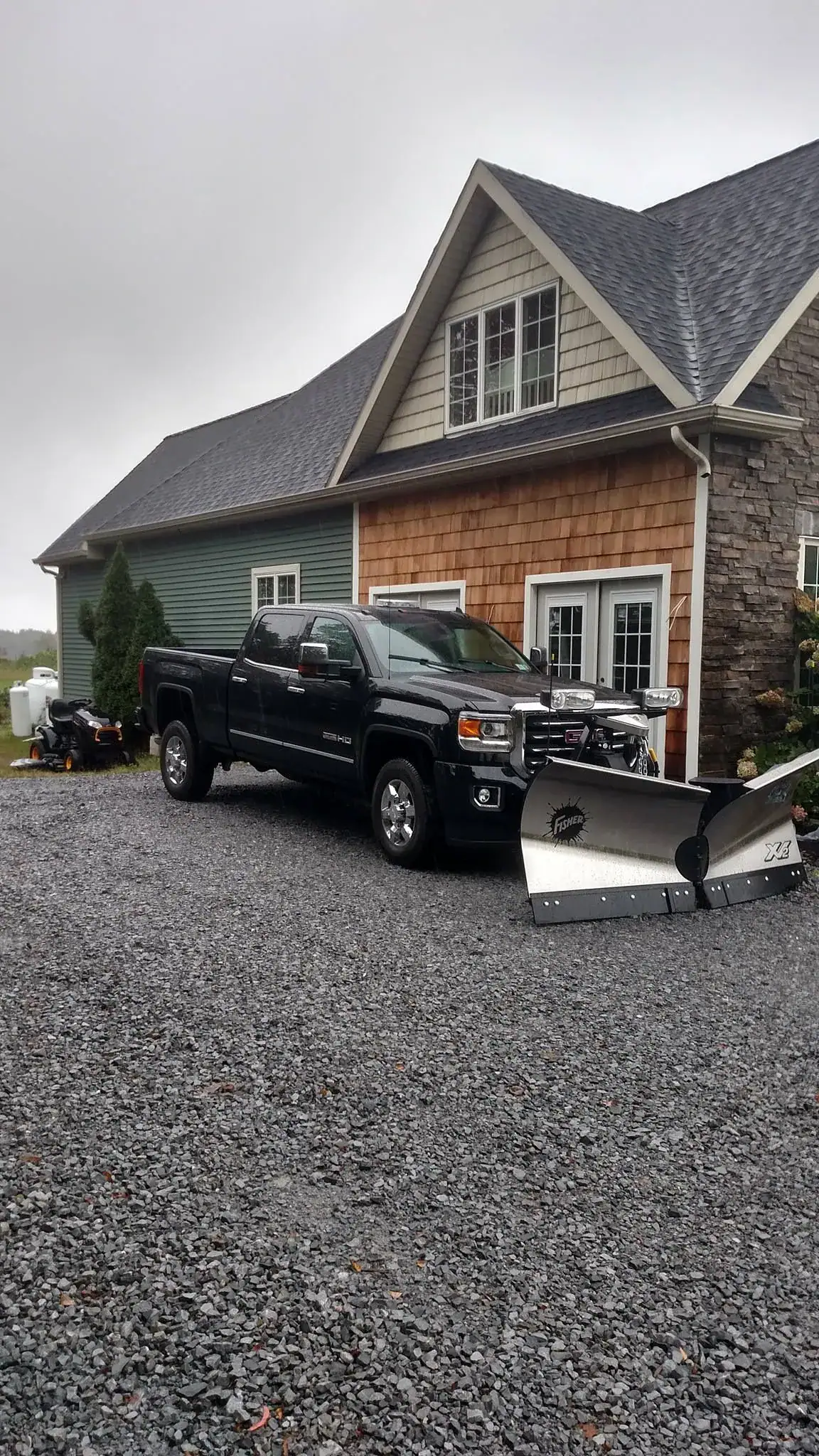 A black pickup truck with a large snow plow attachment—ideal for a paving contractor Saratoga & Warren County, NY—is parked on a gravel driveway in front of a house with green and brown siding and stone accents. A snowmobile is also visible beside the house.