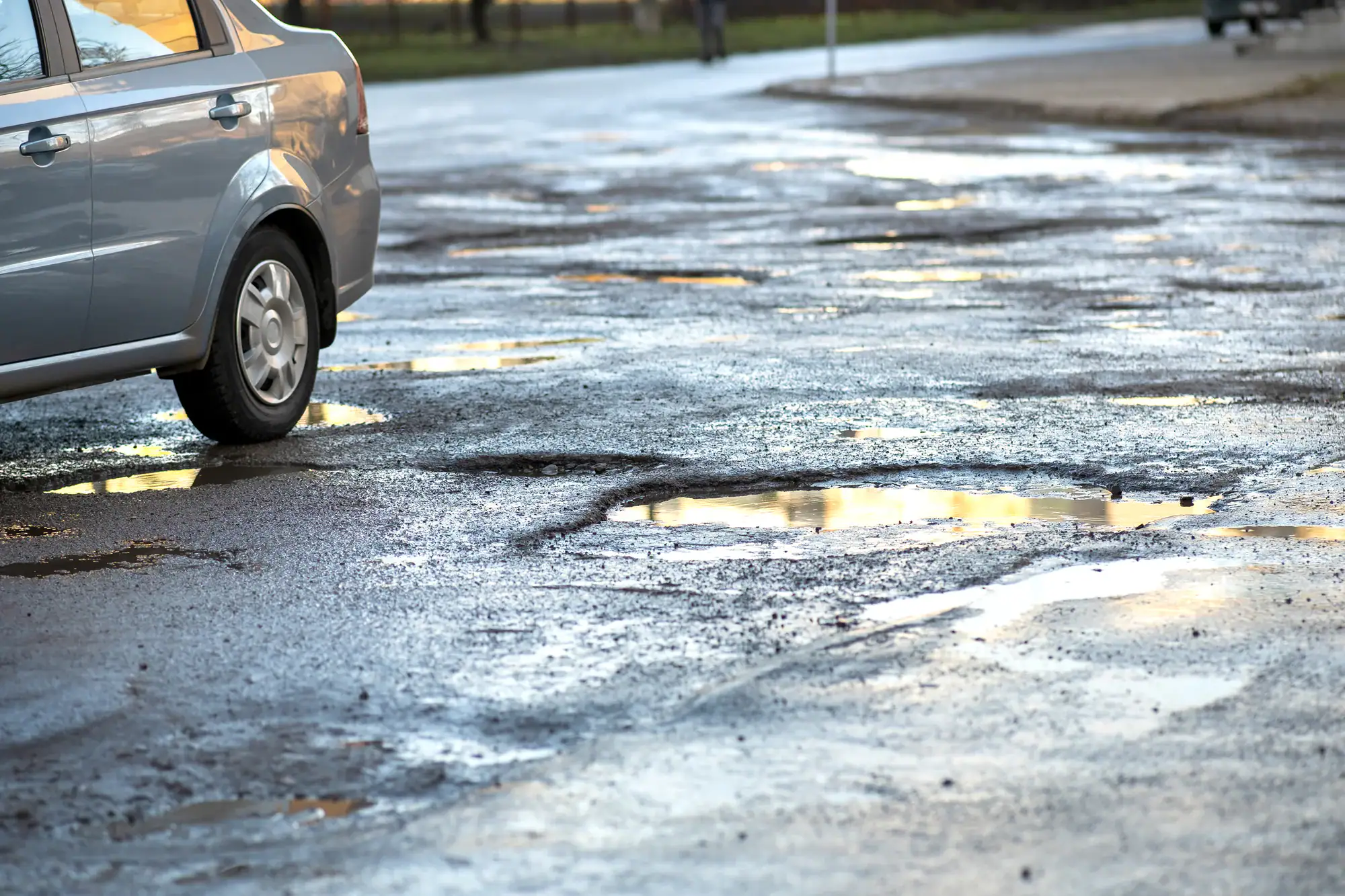 A car is parked on a wet, damaged road with many potholes filled with water, reflecting the sky. The scene suggests poor road conditions and recent rainfall.