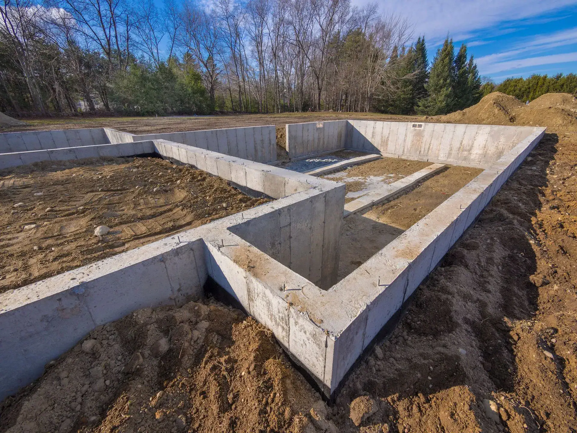Concrete foundation walls form the base outline of a new building on a construction site, surrounded by dirt, trees, and clear blue sky in the background.