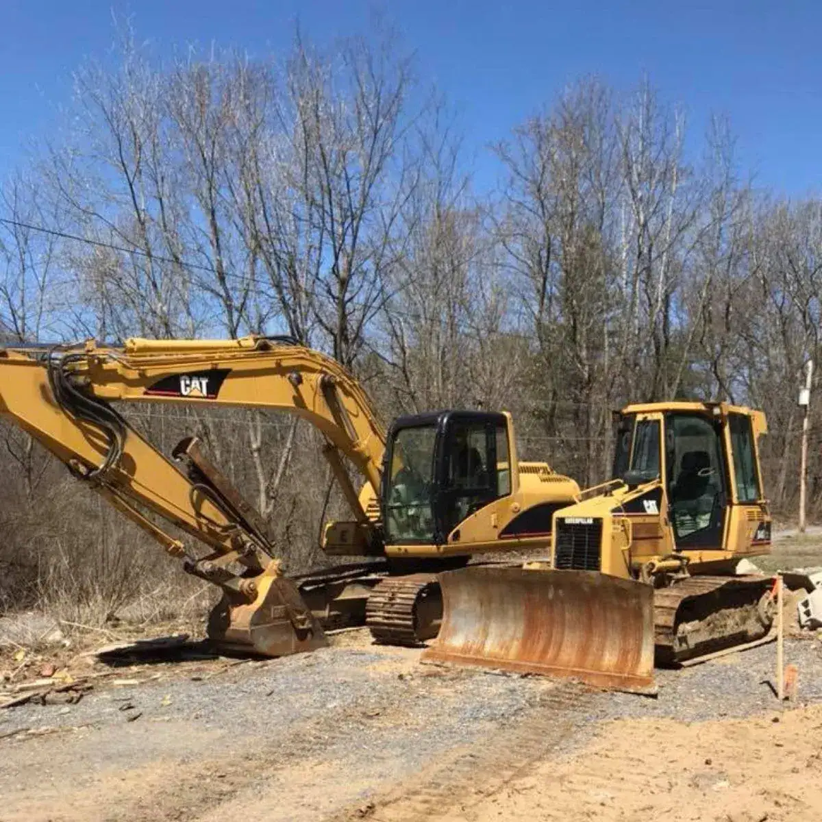 A yellow excavator and a yellow bulldozer are parked closely together at a construction site surrounded by bare trees and dirt under a clear blue sky.