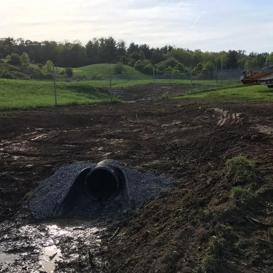 A plastic drainage pipe surrounded by gravel emerges from muddy ground, with tire tracks and a puddle nearby. In the background, grassy fields, trees, a chain-link fence, and construction equipment are visible under a cloudy sky.