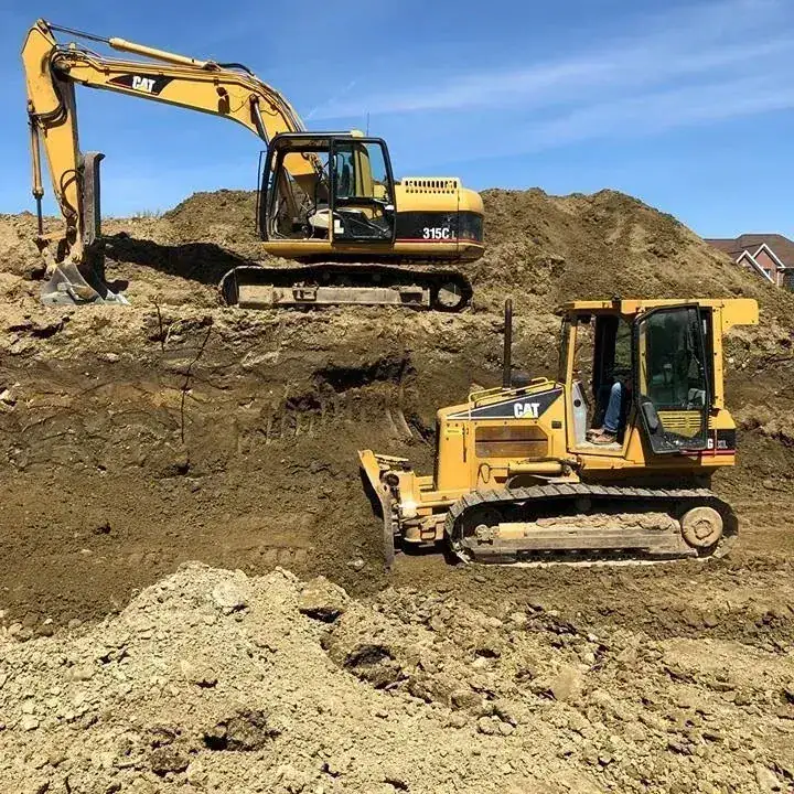 Two yellow CAT construction vehicles, an excavator and a bulldozer, work on a dirt mound at a construction site under a clear blue sky. Houses are visible in the background.