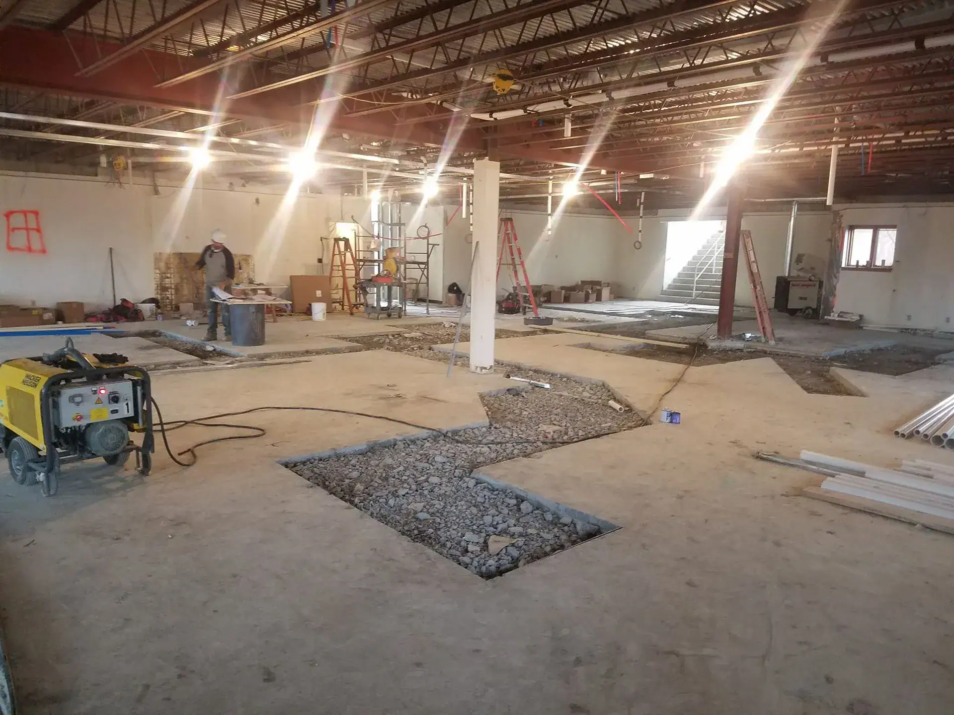 A large indoor construction site with exposed beams, workers, ladders, a generator, tools, and open trenches in the concrete floor, illuminated by bright overhead lights.