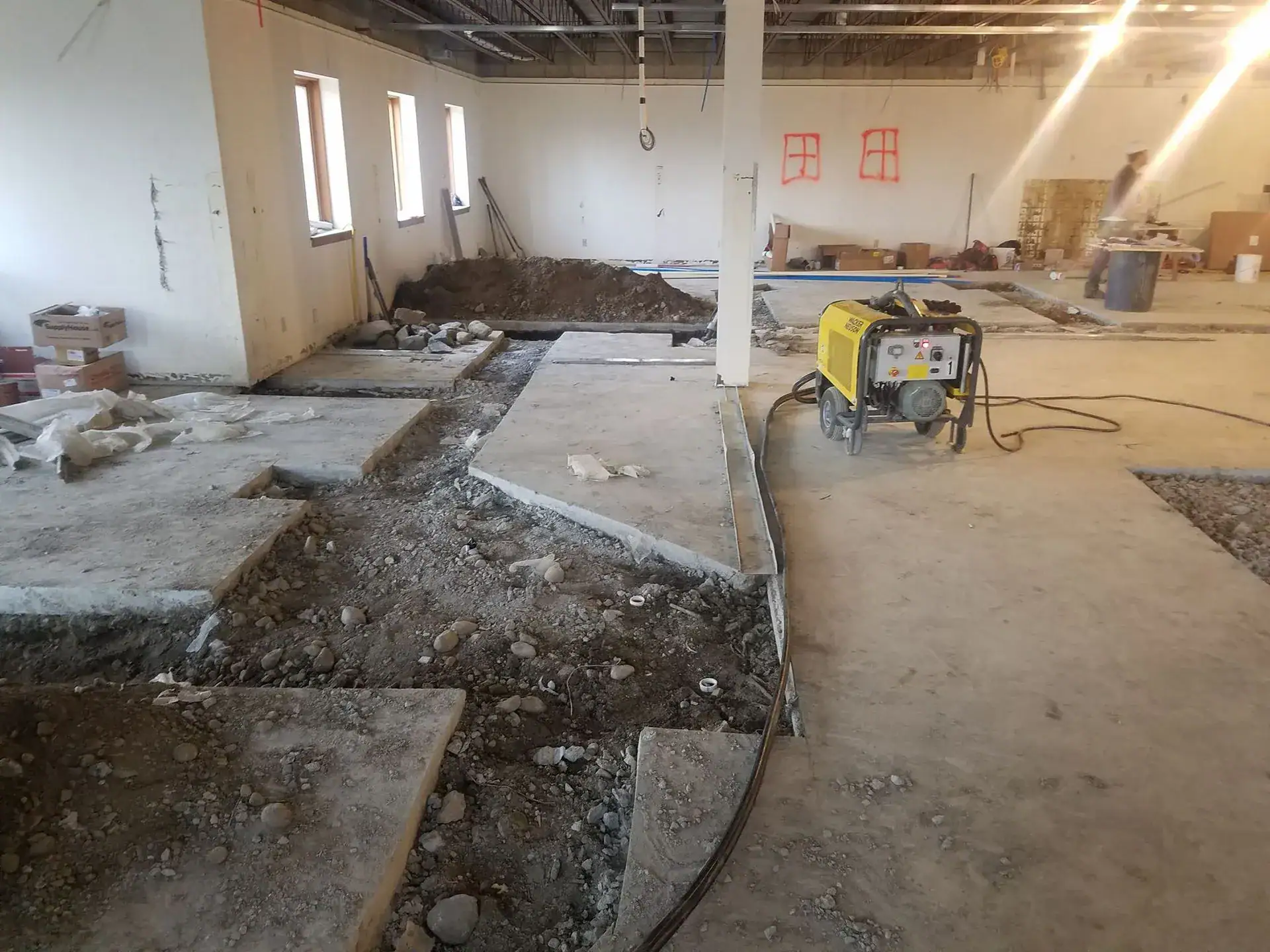 A construction site inside a building shows sections of concrete flooring removed, exposing dirt and rocks. Construction equipment and materials are scattered throughout the space.