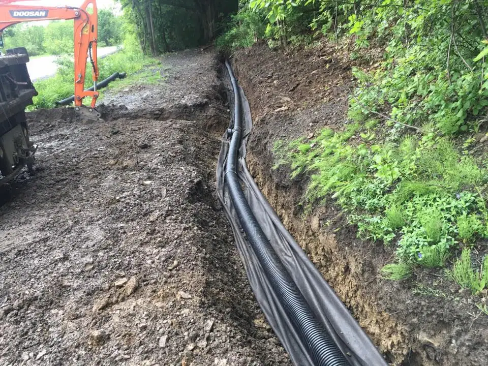 A trench dug alongside a dirt road with black corrugated drainage pipes installed at the bottom, surrounded by soil and green vegetation; an orange excavator is visible in the background.