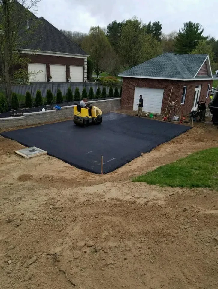 A paving contractor from Saratoga & Warren County, NY operates a yellow steamroller on freshly laid asphalt in a residential driveway, with a brick garage and house in the background. Other workers and construction tools are visible nearby.