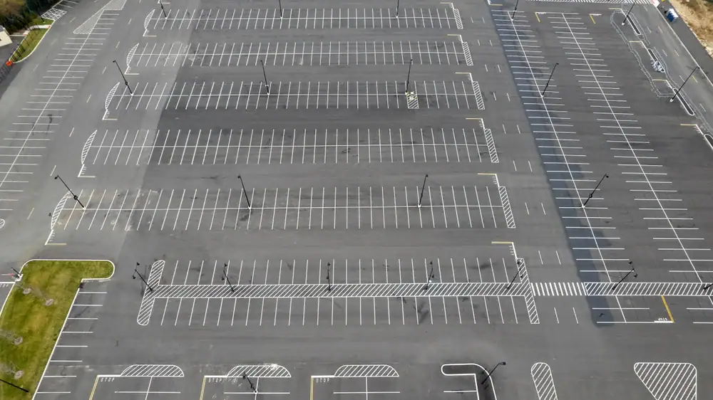 Aerial view of a large, empty parking lot with multiple rows of parking spaces and clearly marked lines and crosswalks. No vehicles are present, and some small patches of grass are visible at the edges.
