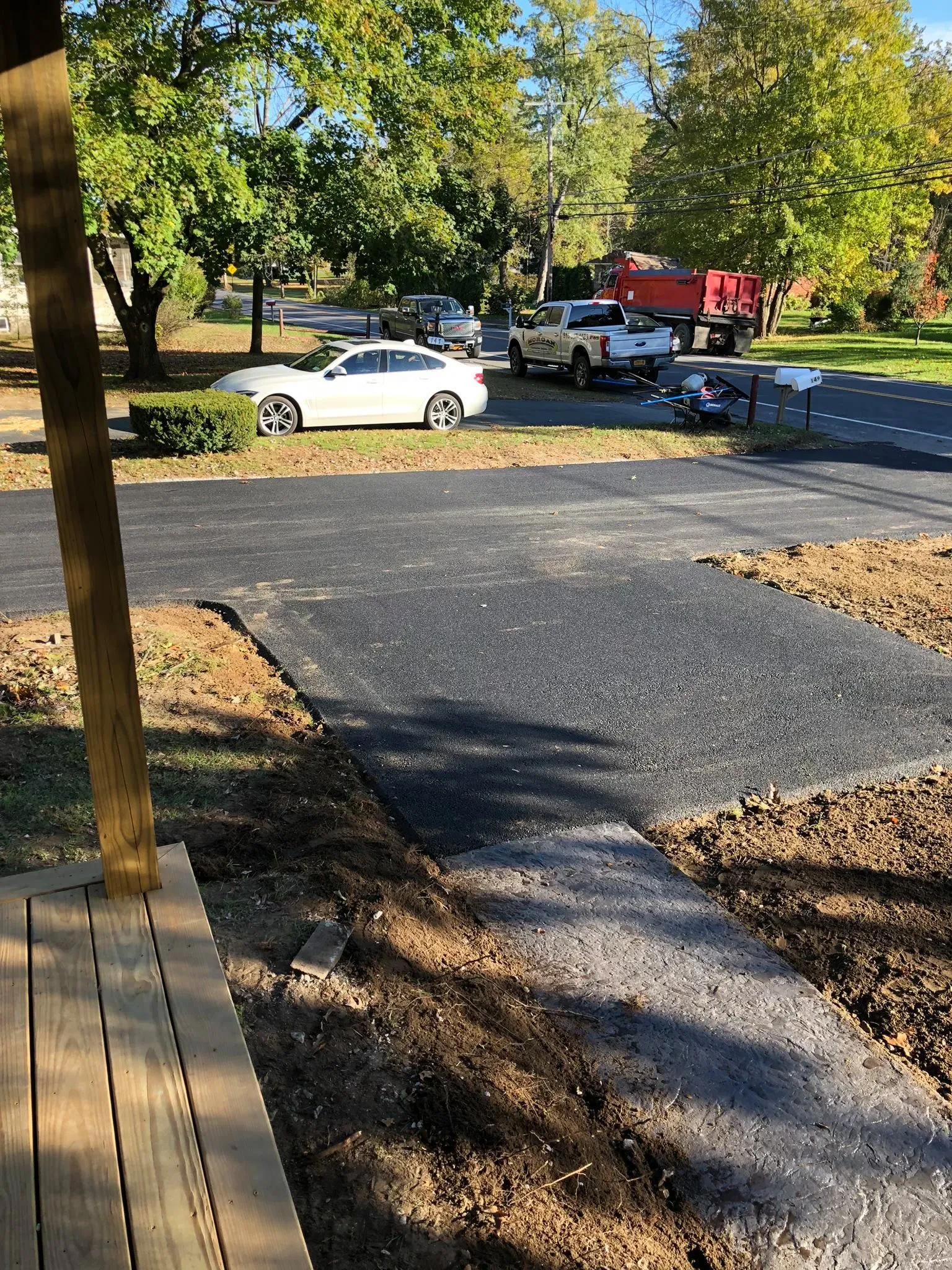 View from a porch showing a newly paved driveway by a paving contractor in Saratoga & Warren County, NY, leading to a suburban street with parked cars and trees with green foliage. A mailbox is visible near the road.