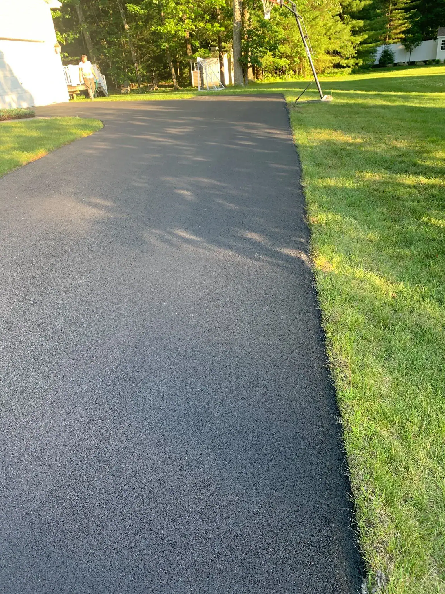 A freshly paved black asphalt driveway curves through a green lawn, with sunlight filtering through trees and a basketball hoop visible in the background.