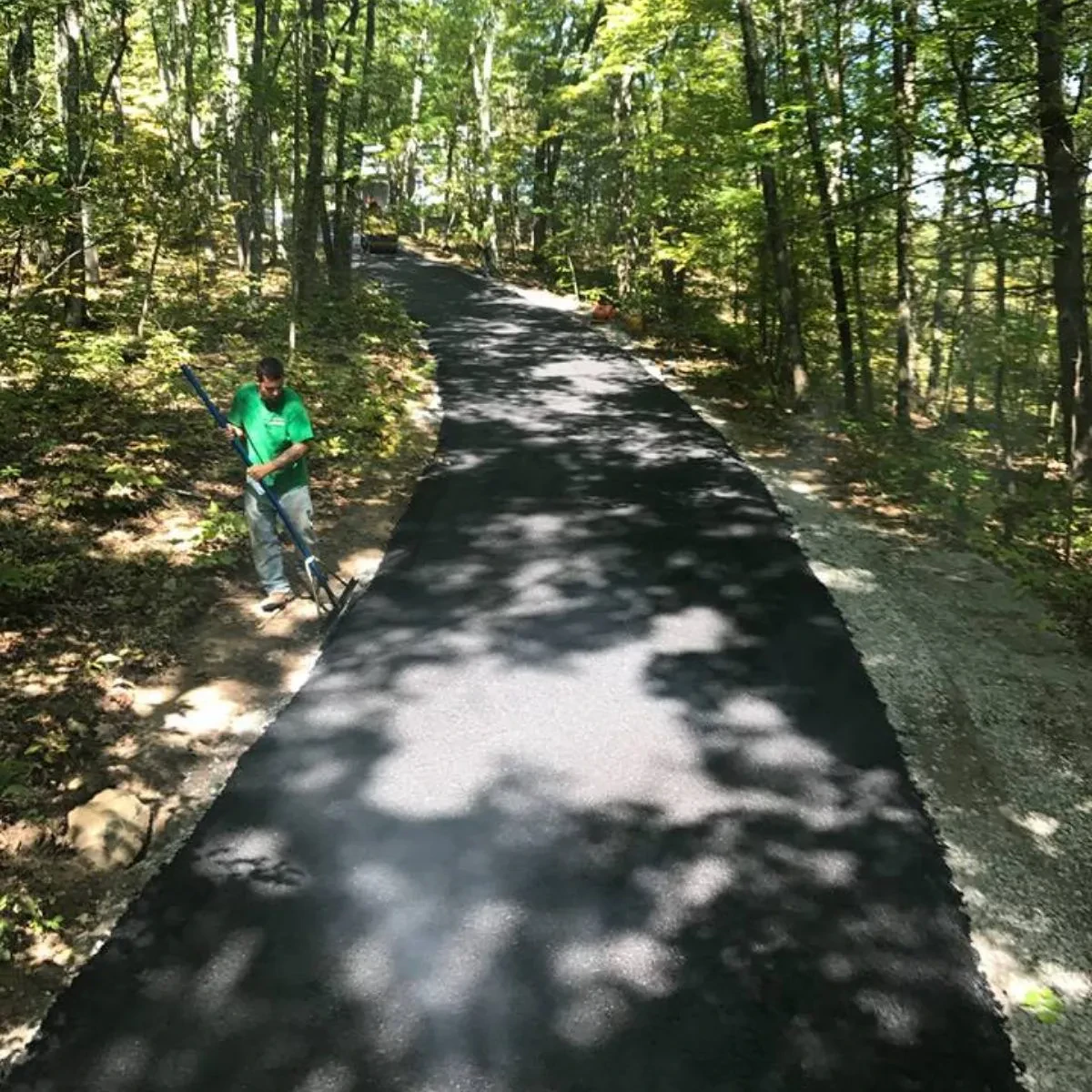 A person in a green shirt stands next to a newly paved black asphalt path, crafted by a paving contractor Saratoga & Warren County, NY, winding through a sunlit, wooded area with tall trees and dappled shade.