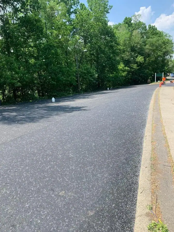 A newly paved road curves to the right, bordered by a sidewalk and lush green trees under a partly cloudy sky. White plastic jugs are spaced out along the edge of the road.