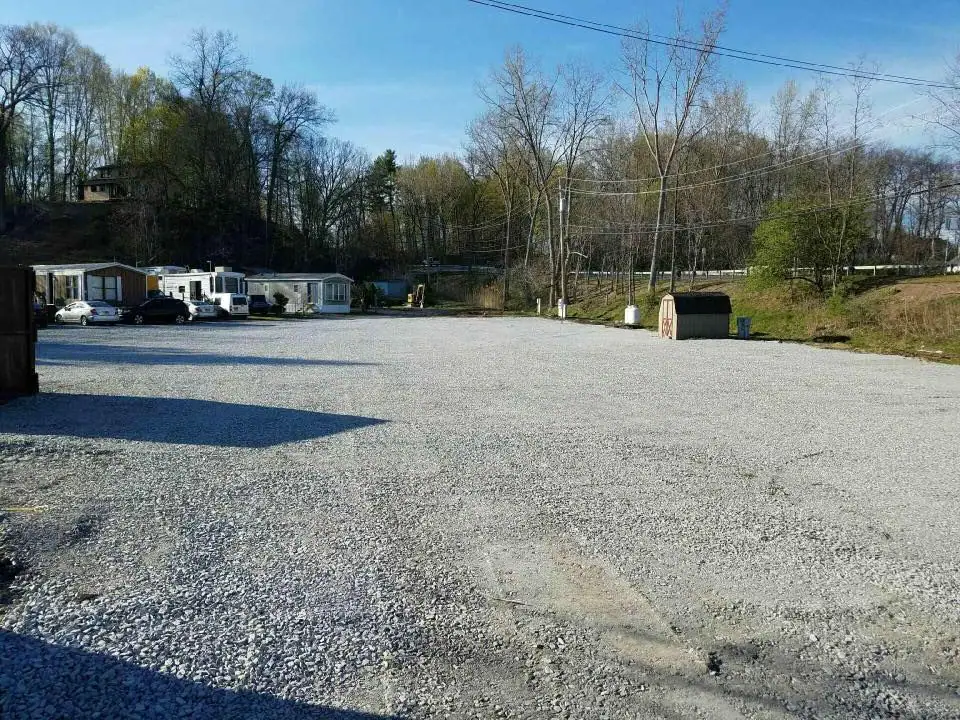 A large gravel parking lot with a few mobile homes and trees in the background on a sunny day. A small dumpster and utility poles are visible near the lot’s edge.