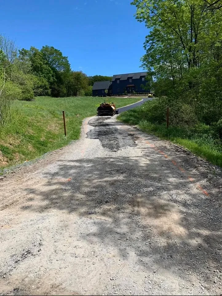 A gravel driveway leads uphill toward a house surrounded by trees and grass in NY, with a truck carrying rolled sod partway up the road under a clear blue sky&mdash;ideal for any paving contractor Saratoga & Warren County might recommend.