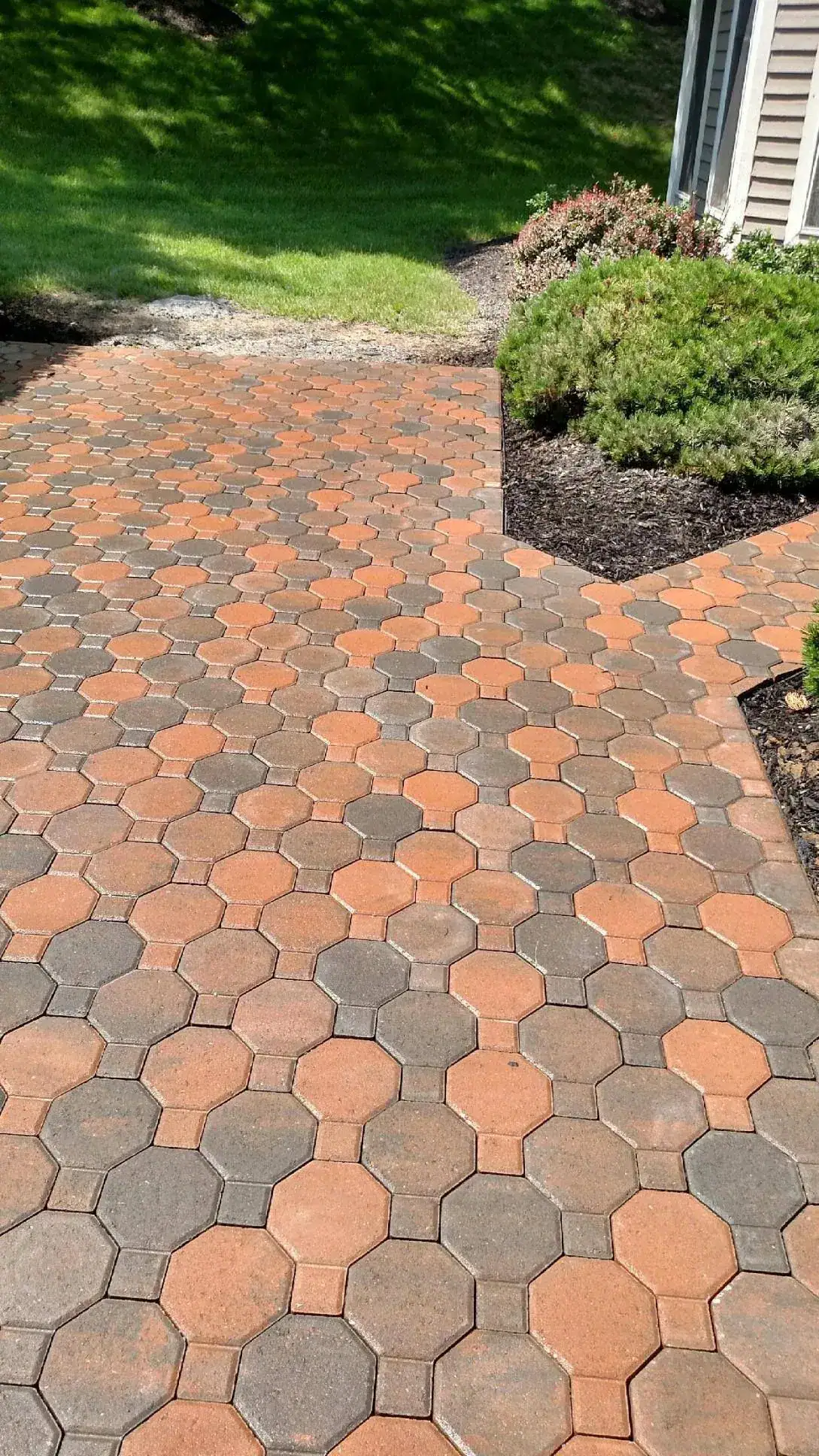 A walkway made of interlocking hexagonal and square pavers in shades of brown and tan leads through a landscaped yard with green grass, bushes, and a beige house.