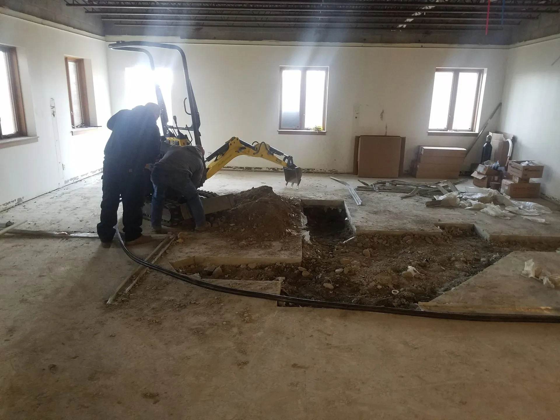 Two people, working for a paving contractor in Saratoga & Warren County, NY, operate a small excavator indoors, digging up concrete flooring. Loose dirt and construction debris are scattered around the partially demolished room.