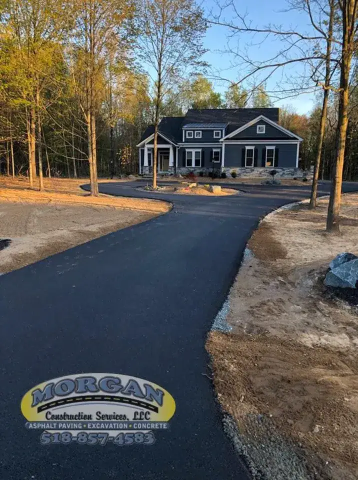 A newly paved asphalt driveway curves through a wooded area, leading to a gray house with white trim. The Morgan Construction Services, LLC logo and contact number are visible in the bottom left corner.
