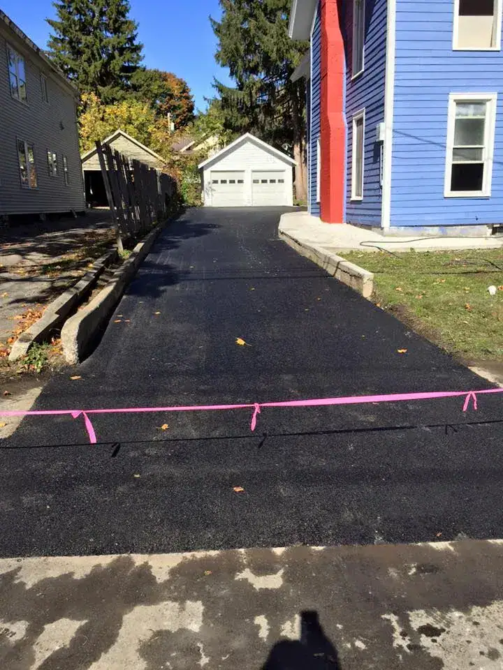 A newly paved black asphalt driveway leads to a garage behind a blue house, with a pink caution tape across the entrance. Trees with autumn leaves are visible in the background.