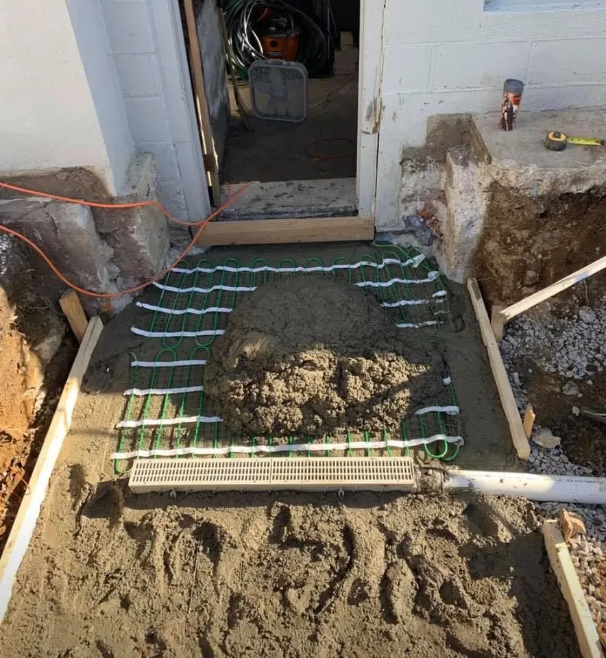 A concrete pad is being poured by a paving contractor Saratoga & Warren County over green heating cables laid on the ground outside a NY building entrance, with tools, cords, and construction materials visible nearby.