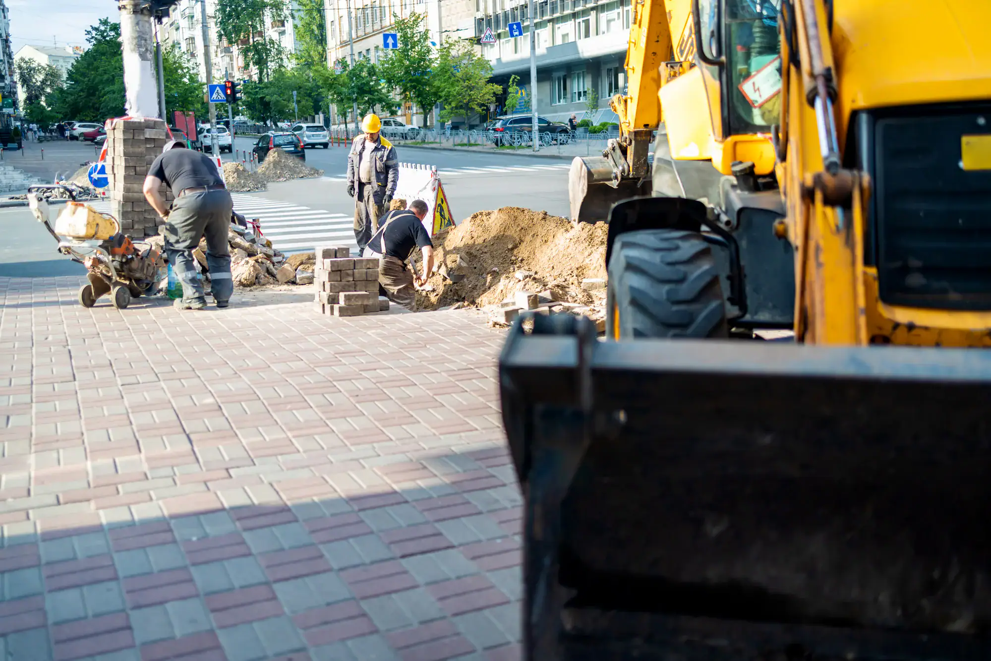 Several construction workers are repairing a sidewalk with paving bricks; a yellow construction vehicle and piles of dirt are visible, with urban buildings and a street in the background.