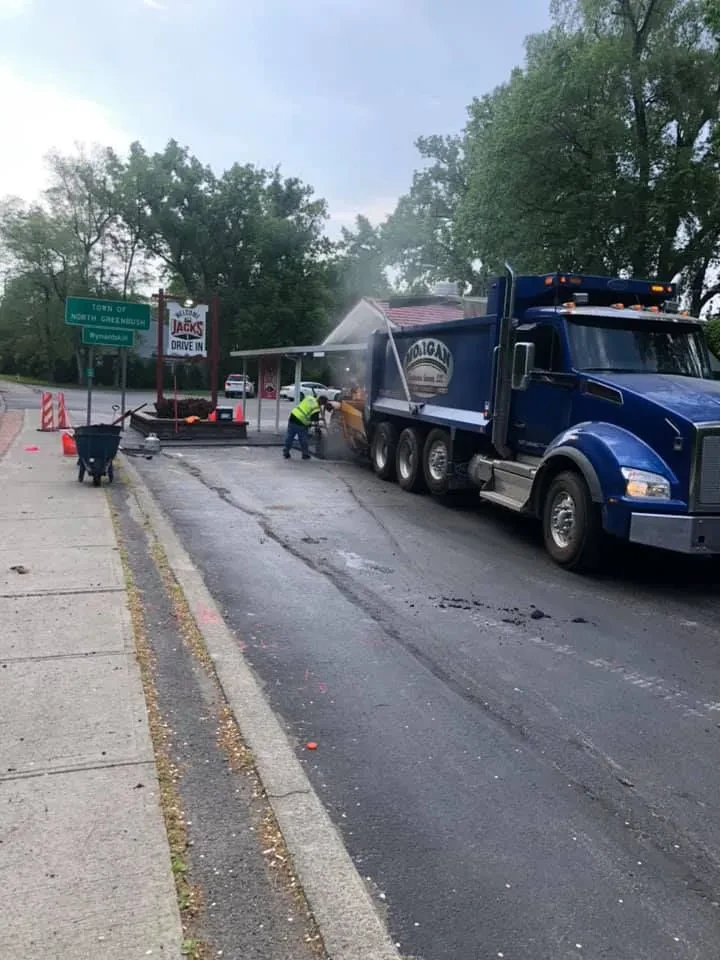 Two workers from a paving contractor Saratoga & Warren County, NY, are resurfacing a road with asphalt from a blue dump truck. Construction cones and signs are nearby, trees line the background, and a "Jack's Drive In" restaurant sign is visible.