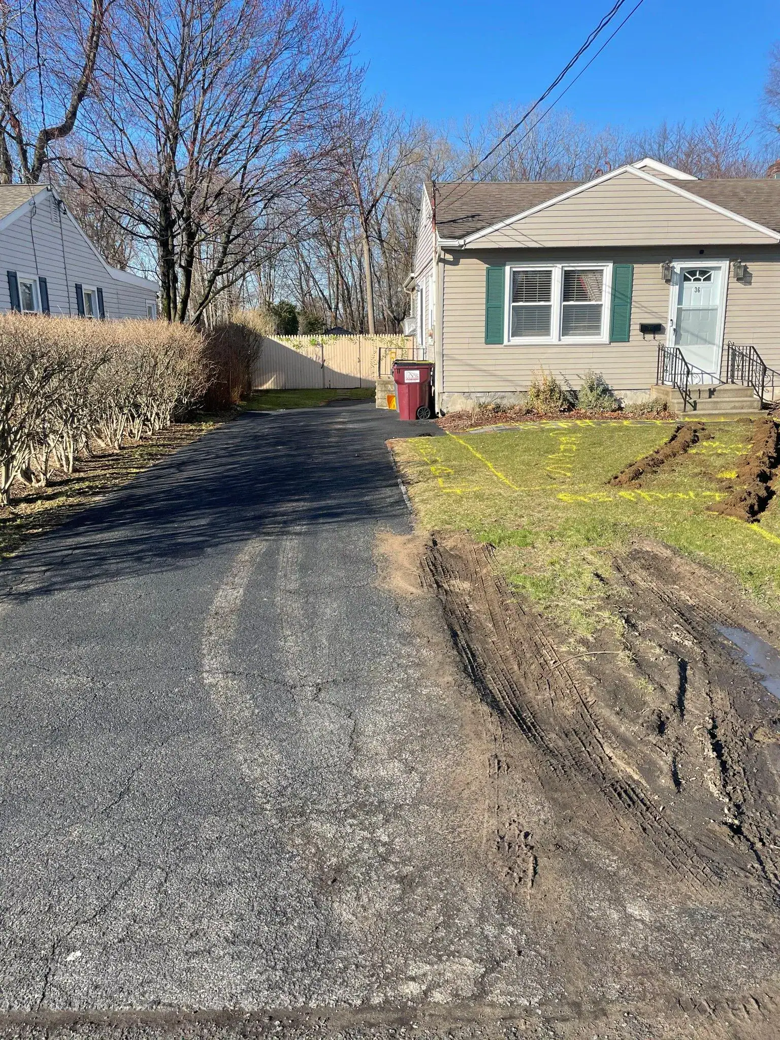 A suburban house with a long driveway, fresh tire tracks in the muddy grass, and trimmed bushes on one side. The sky is clear and blue, with bare trees in the background.