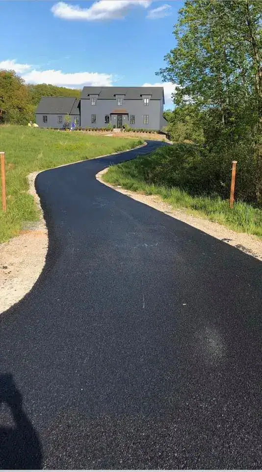 A newly paved black asphalt driveway curves through green grass toward a modern gray house surrounded by trees under a partly cloudy sky.
