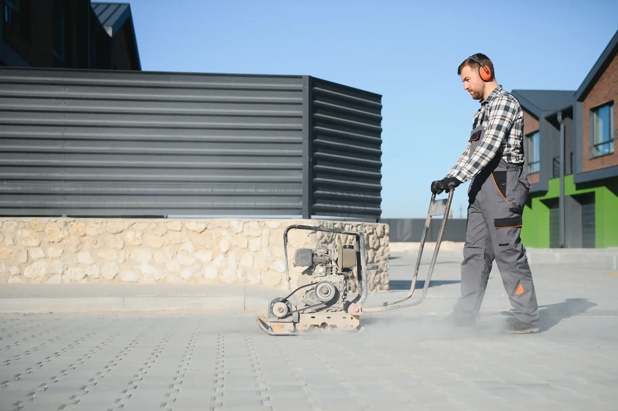 A construction worker wearing ear protection and gloves operates a vibrating plate compactor on a paved outdoor surface, with dust rising around the machine on a sunny day near modern buildings.