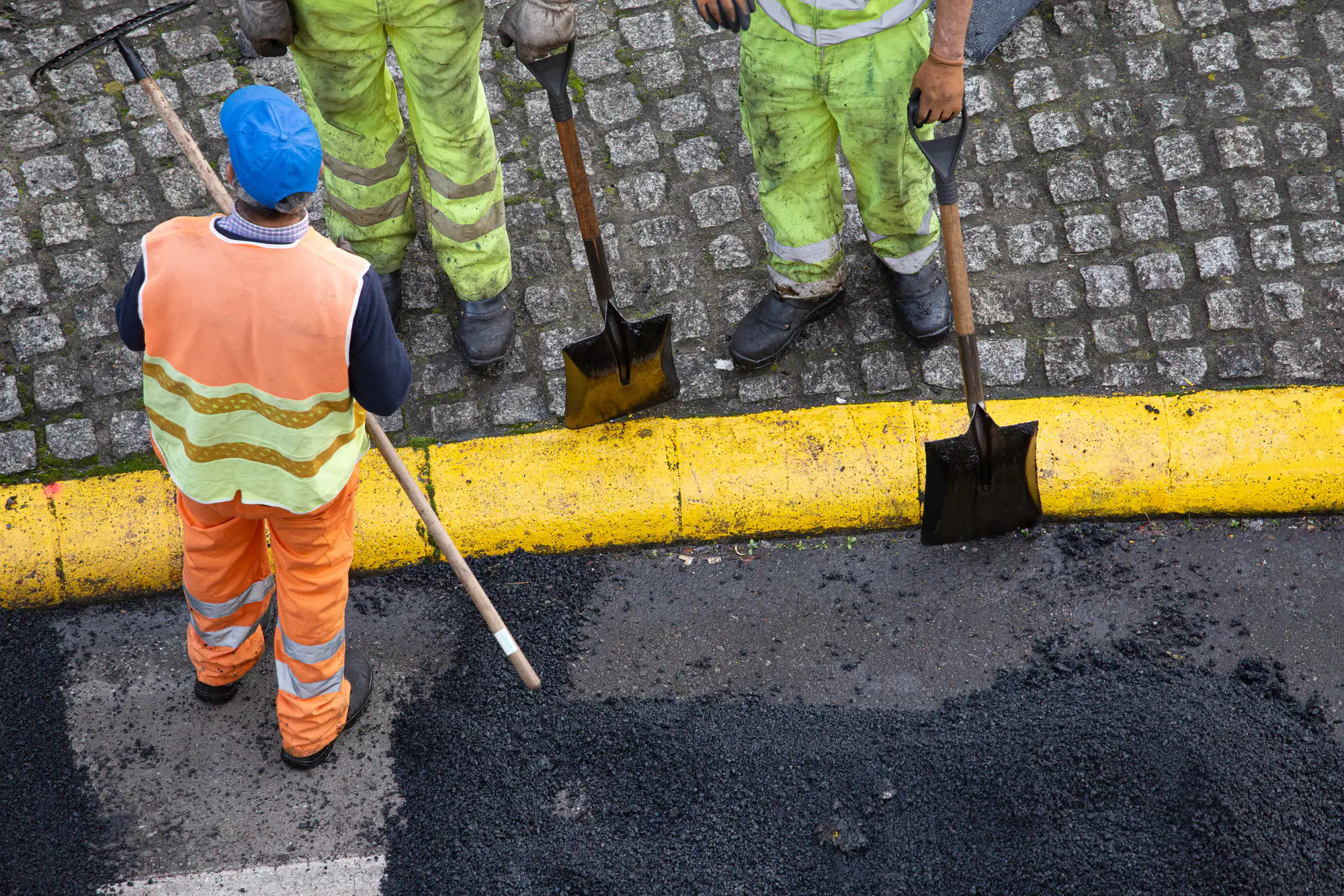 Aerial view of three road workers in reflective clothing standing on cobblestone beside a yellow curb, holding shovels, with one spreading asphalt on the street.