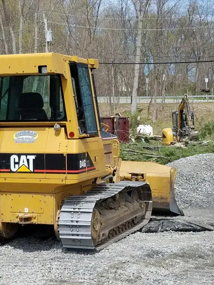 A yellow CAT D4G bulldozer is parked on gravel at a construction site, with another construction vehicle and trees visible in the background.